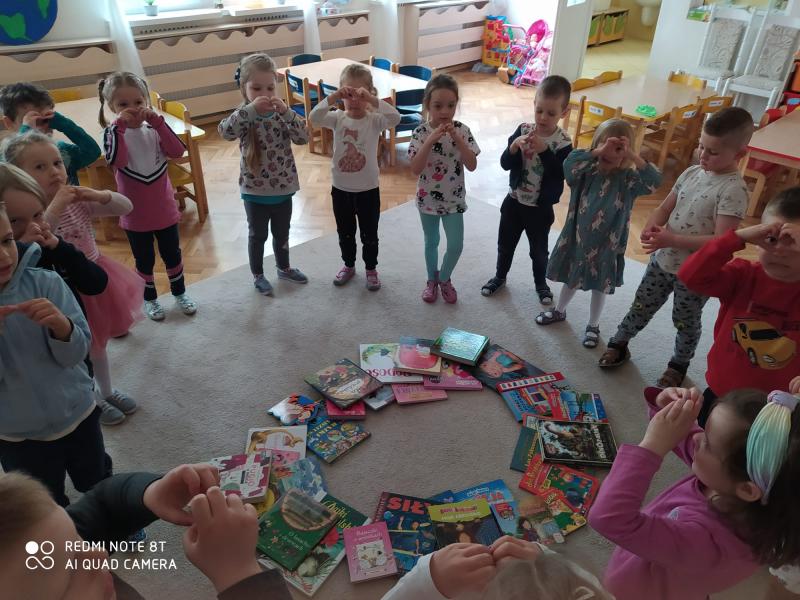 Dzieci stoją wokół książek rozłożonych na dywanie.