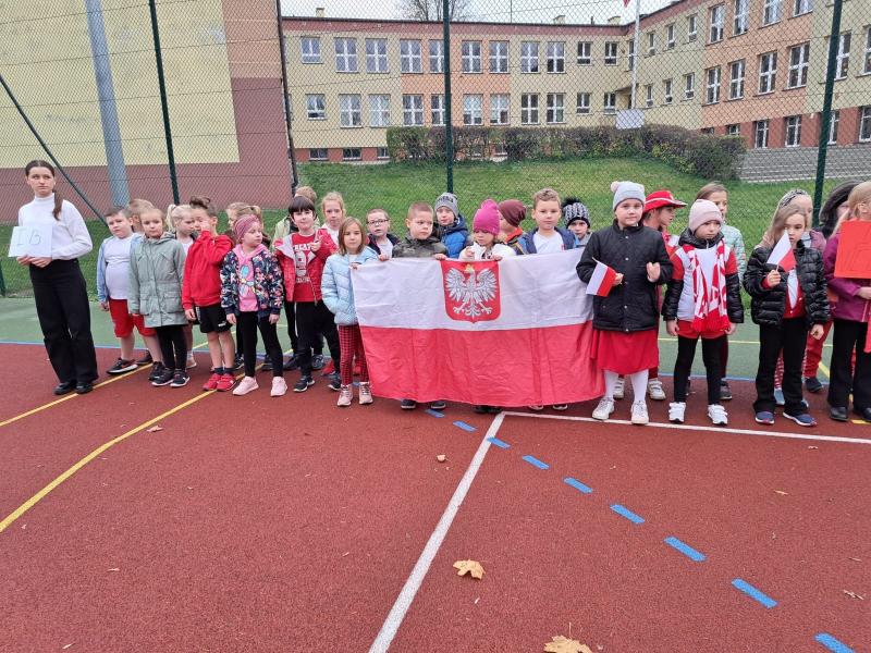 Kibice z młodszych klas stoją na boisku szkolnym, trzymają flagę narodową.