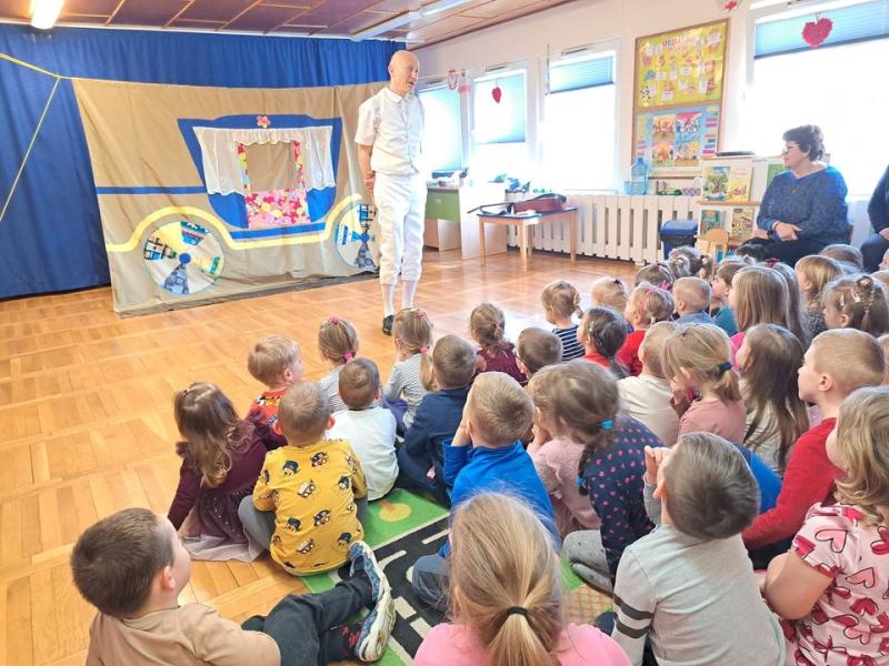Scenografia - kolorowa Karoca wyszyta na wielkim płótnie, przed nią na scenie mężczyzna ubrany na biało, jego twarz skierowana do widowni - dzieci siedzących w rzędach odwróconych tyłem