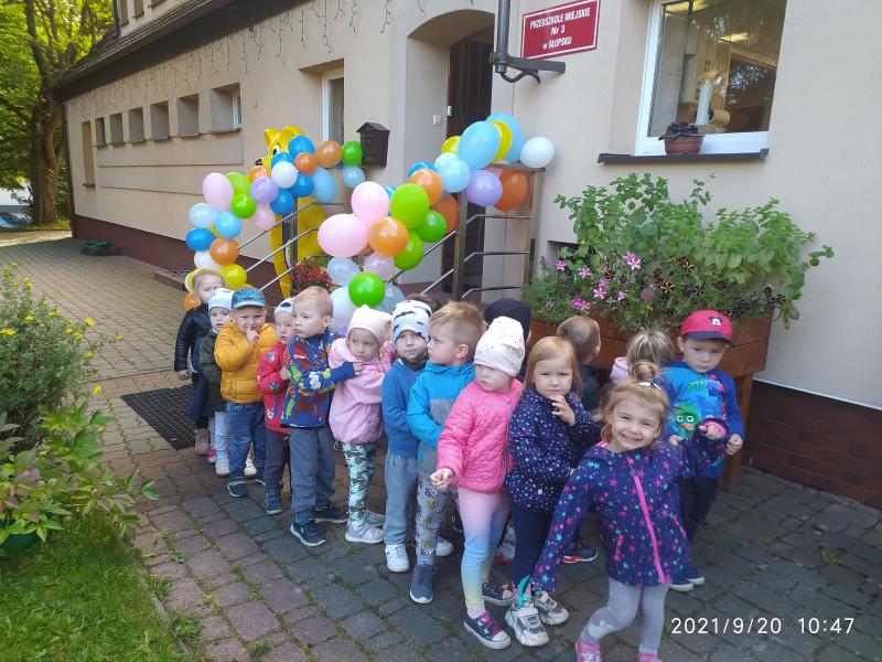 Dzieci ustawione jeden za drugim przy wyjściu z przedszkola udekorowanego balonami