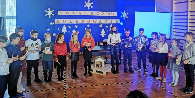 Scena sali gimnastycznej. Na zdjęciu uczniowie klas 1-4 śpiewające kolędy, małymi instrumentami w dłoniach. W tle błękitna dekoracja świąteczna.