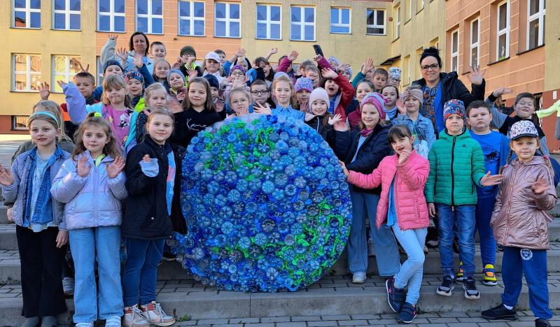 Uczniowie na placu apelowym szkoły prezentują wielką kulę ziemską wykonaną z niebieskich i zielonych plastikowych butelek.