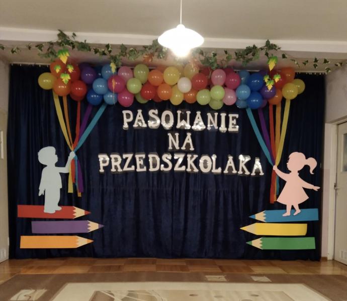 Dekoracja - granatowa zasłona a na niej biały napis "pasowanie na przedszkolaka" balony, postać chłopca i dziewczynki stojących na kredkach