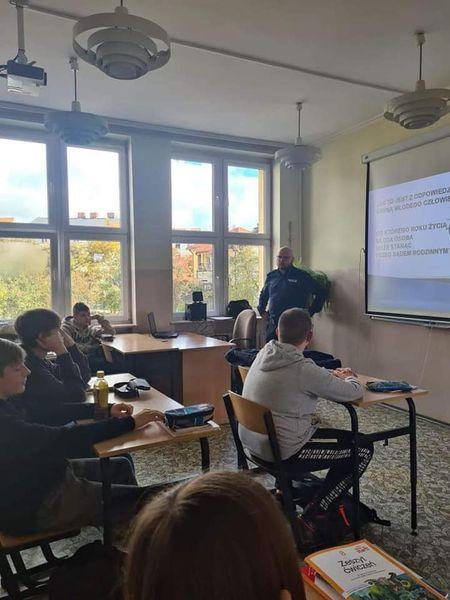 Gabinet lekcyjny pełen uczniów. Przy tablicy multimedialnej z prezentacją stoi policjant