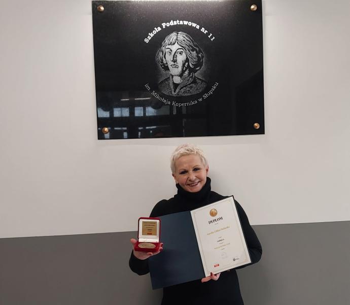 Nauczyciel roku Pani Aurelia Gibka-Pukiełko z dyplomem i medalem. W tle tablica pamiątkowa szkoły.