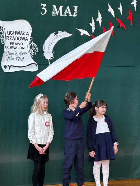 Uczniowie na scenie sali gimnastycznej z strojach galowych z flagą Polski