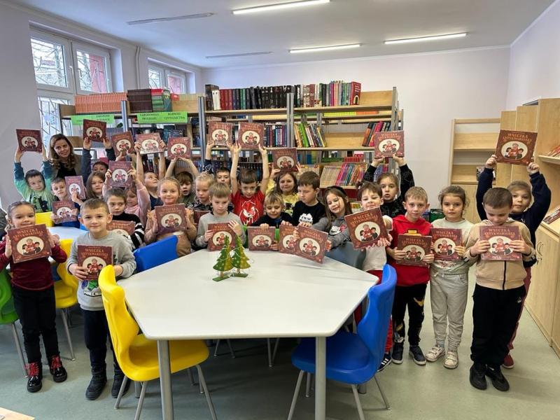 Zdjęcie grupowe klasy pierwszej z nauczycielem bibliotekarzem w szkolnej bibliotece. Uczniowie pokazują otrzymaną książkę.