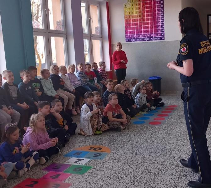 Uczniowie klas 2-3 na szkolnym holu słuchają wykładu strażniczki miejskiej