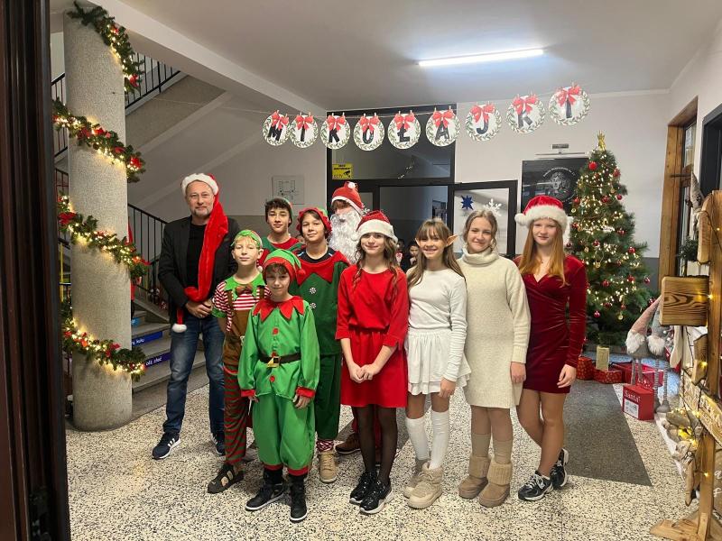 Zdjęcie uczniów Samorządu Uczniowskiego w świątecznych strojach. Po lewej stronie Dyrektor Szkoły.  W tle choinka i świąteczne dekoracje na holu szkoły.