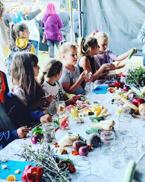 Dzieci w ferworze kiszenia. Siedzą przy stole z warzywami i owocami. Obierają i kroją produkty, które potem układają w słoikach.