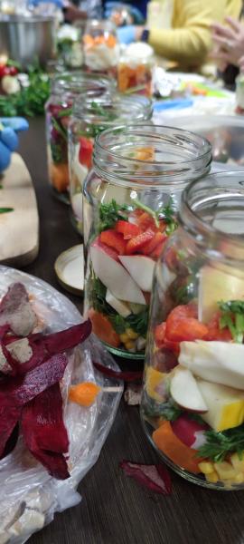 Na stole stoją słoiki częściowo wypełnione warzywami oraz owocami. W tle widać ręce uczestników warsztatów, obok słoików obierki buraków.