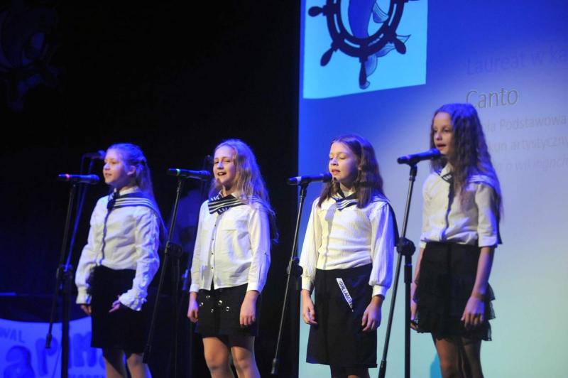 Dziewczynki ubrane w stroje marynarskie, stoją na scenie przy mikrofonach