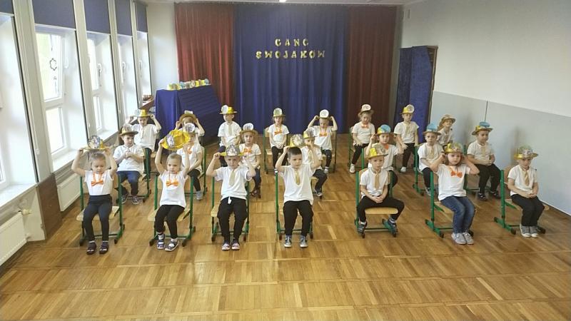 Dzieci siedzą na krzesełkach trzymając rekwizyty – kapelusze z postaciami Swojaków