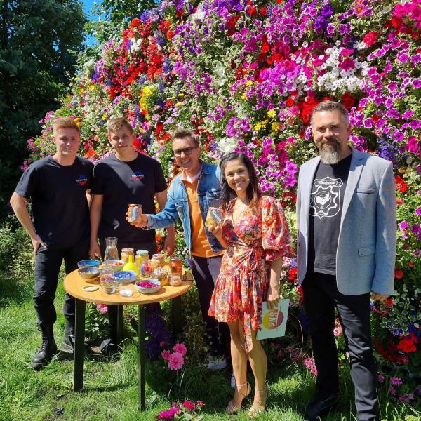Uczniowie Maksymilian i Marcin stoją w ogrodzie TVP z prowadzącymi Pytania Na Śniadanie Robertem i Katarzyną oraz nauczycielem Marcinem pozują do zdjęcia przy stole z kiszonkami oraz kwiatami. W tle ściana kolorowych kwiatów.
