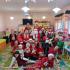 Dzieci ubrane na czerwono siedzą i trzymają prezenty widać Mikołaja i Panie Mikołajowe.
