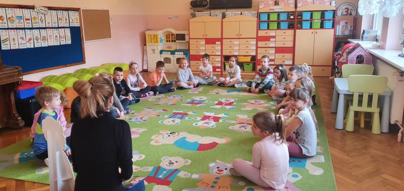 Dzieci siedzą na dywanie w kole i słuchają książeczki czytanej przez Panią Martą Kotusiewicz