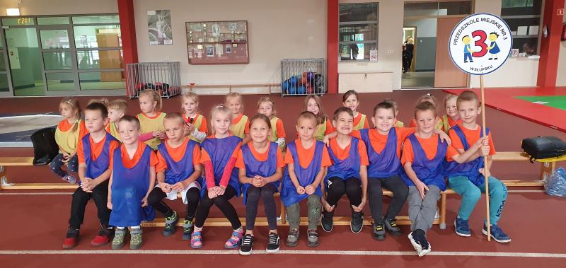 dzieci w pomarańczowych koszulkach i niebieskich kamizelkach siedzą na ławeczce , jeden chłopiec trzyma logo przedszkola