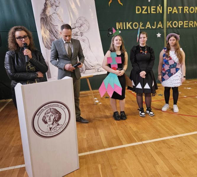 Dyrektor i nauczyciel matematyki, wręczają nagrody i wyróżnienia w konkursie matematyczno-sportowym związanym z liczbą Pi uczennicom z Ukrainy. Zdjęcie wykonano na sali gimnastycznej podczas apelu z okazji dnia patrona.