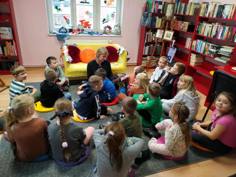Uczniowie klasy 1a w Miejskiej Bibliotece Publicznej Filia nr. 7 w Słupsku. Uczniowie siedzą na podłodze słuchając opowieści bibliotekarki.