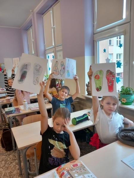 Uczniowie siedzący w ławkach podnoszą do góry karty pracy z obrazkiem kolorowych stóp